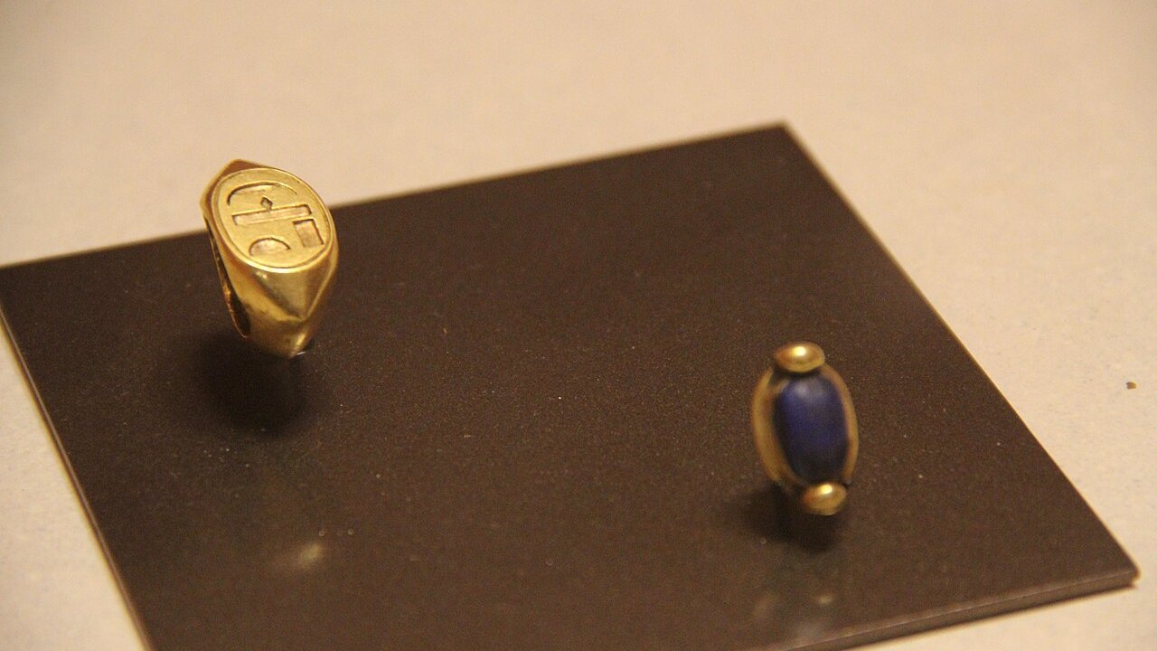 A gold signet ring with an engraved face and a blue lapis-lazuli bead on a dark display tile