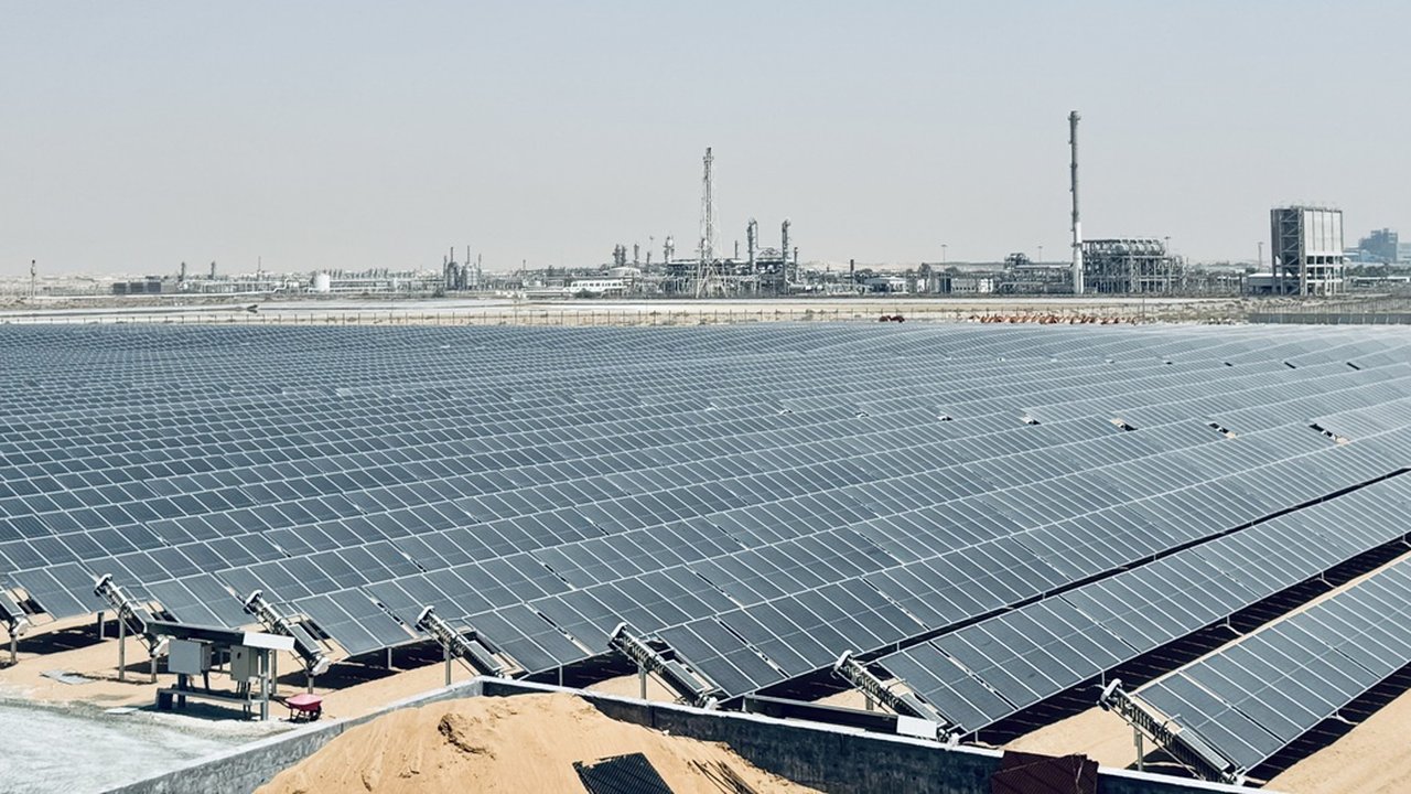 A large field of solar panels on tracking frames in a desert setting, with industrial towers in the distance.