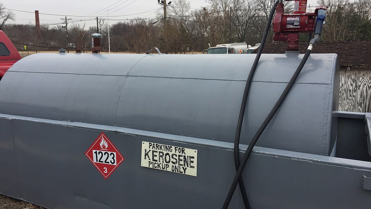A grey industrial fuel storage tank with a hose and pump, labelled as a kerosene pickup point with a hazard placard.