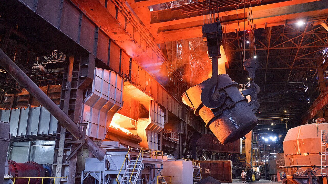 Molten steel being poured from a ladle inside a steelworks with overhead crane and furnace glow