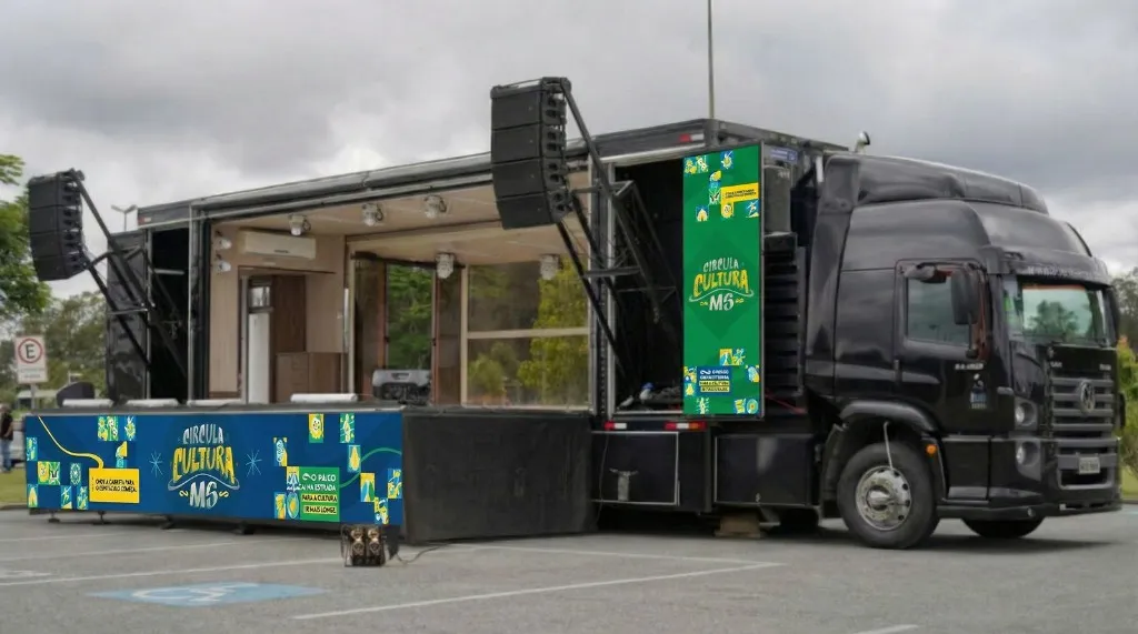 Carreta palco do Circula Cultura MS totalmente aberta e montada para apresentação. A lateral direita se expandiu formando um palco coberto e com piso de madeira, caixas de som profissionais (line array) estão suspensas nas laterais, e equipamentos de iluminação estão instalados no teto.
