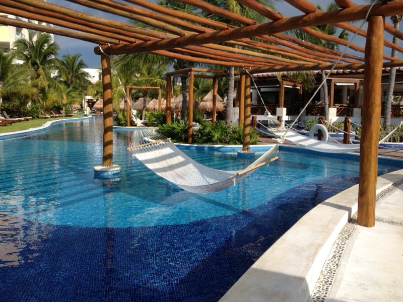 A luxurious resort pool featuring a white hammock suspended over the water beneath a wooden pergola at Excellence Playa Mujeres.