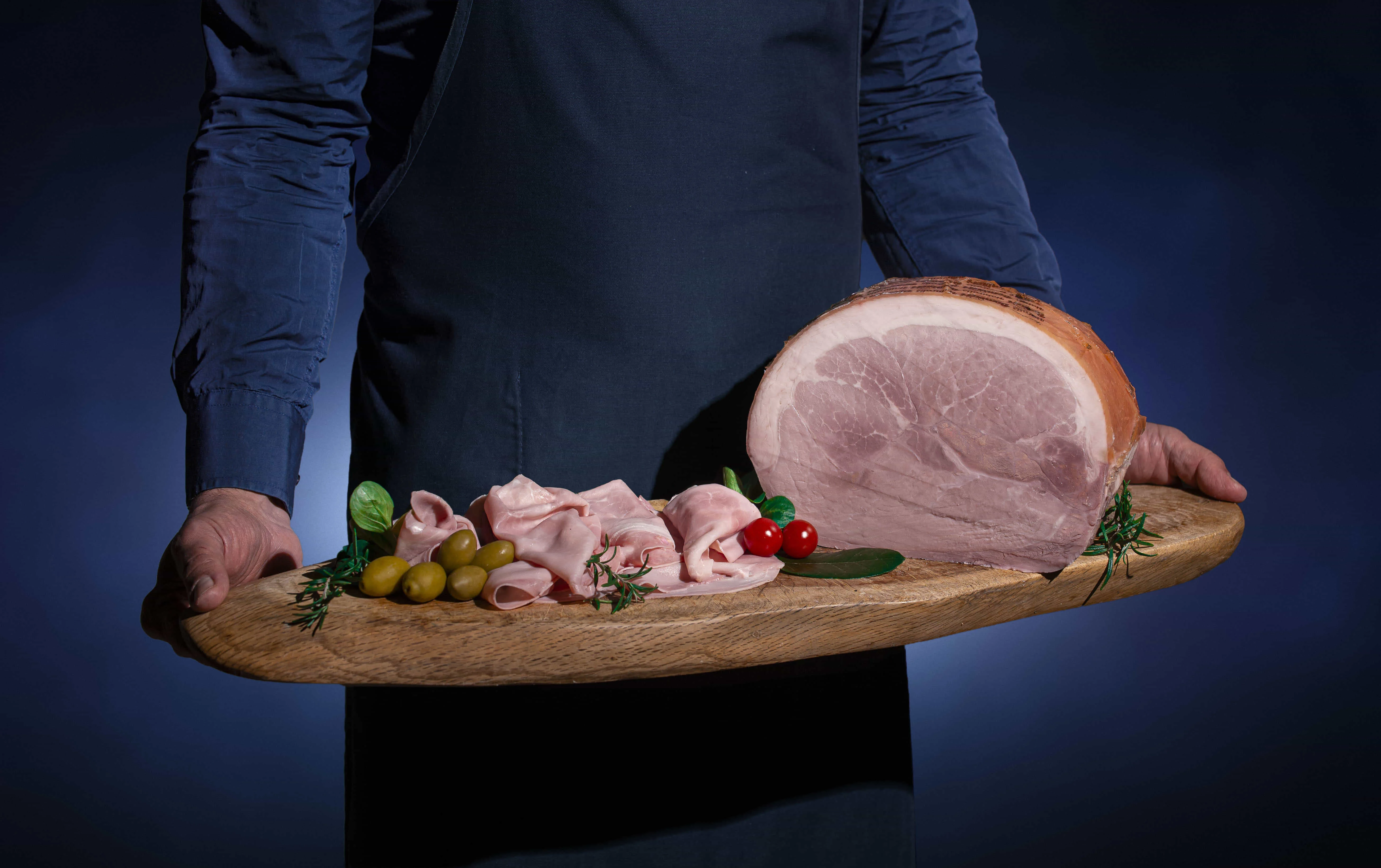 Persona a mezzo busto che tiene in mano un tagliere con sopra il prosciutto cotto su sfondo blu