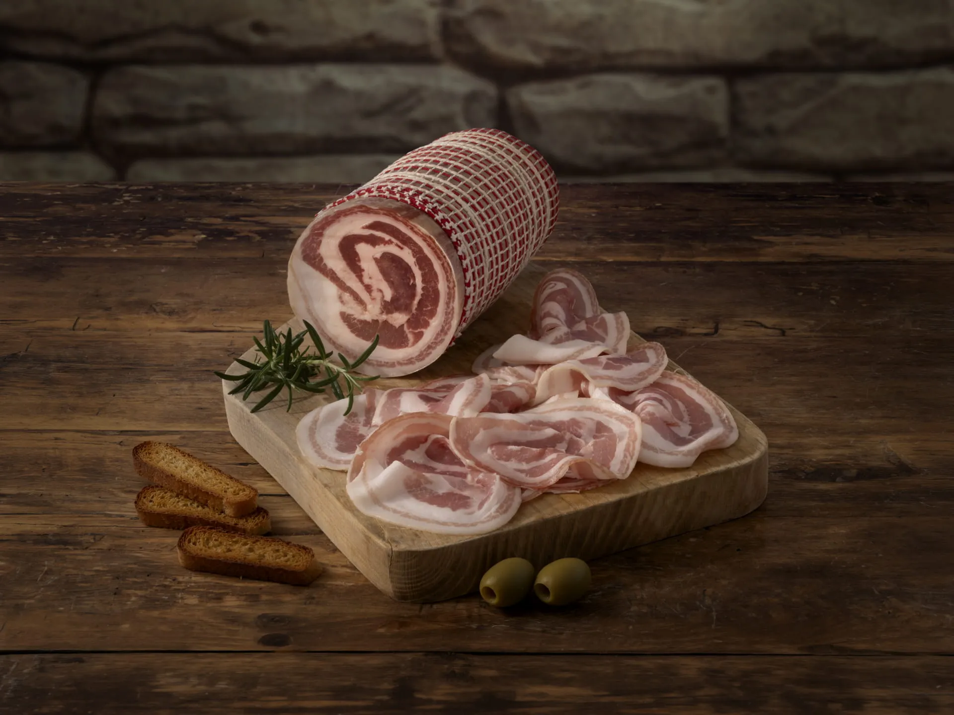 Bombieri rolled and sliced Pancetta arranged on a cutting board with rosemary, croutons, and olives on the side