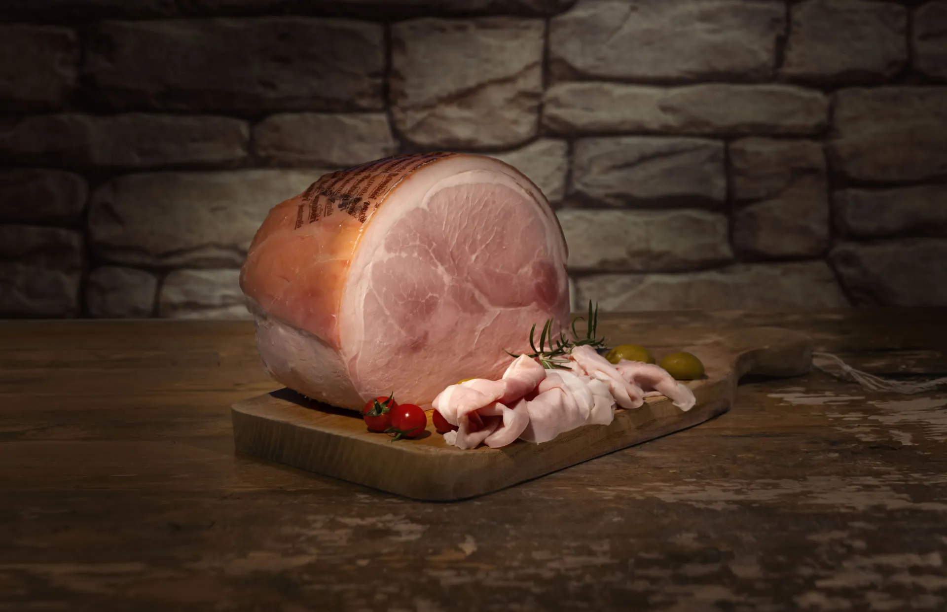 Foto di un prosciutto cotto Bombieri disposto su un tagliere. Accanto il prosciutto affettato con pomodorini e olive