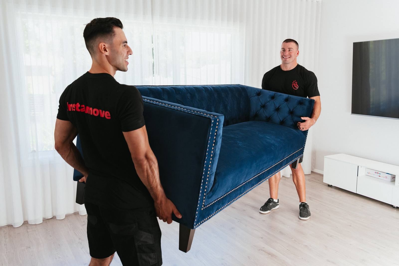 Two movers carry a sofa through a family home during move-in, with packed boxes visible.