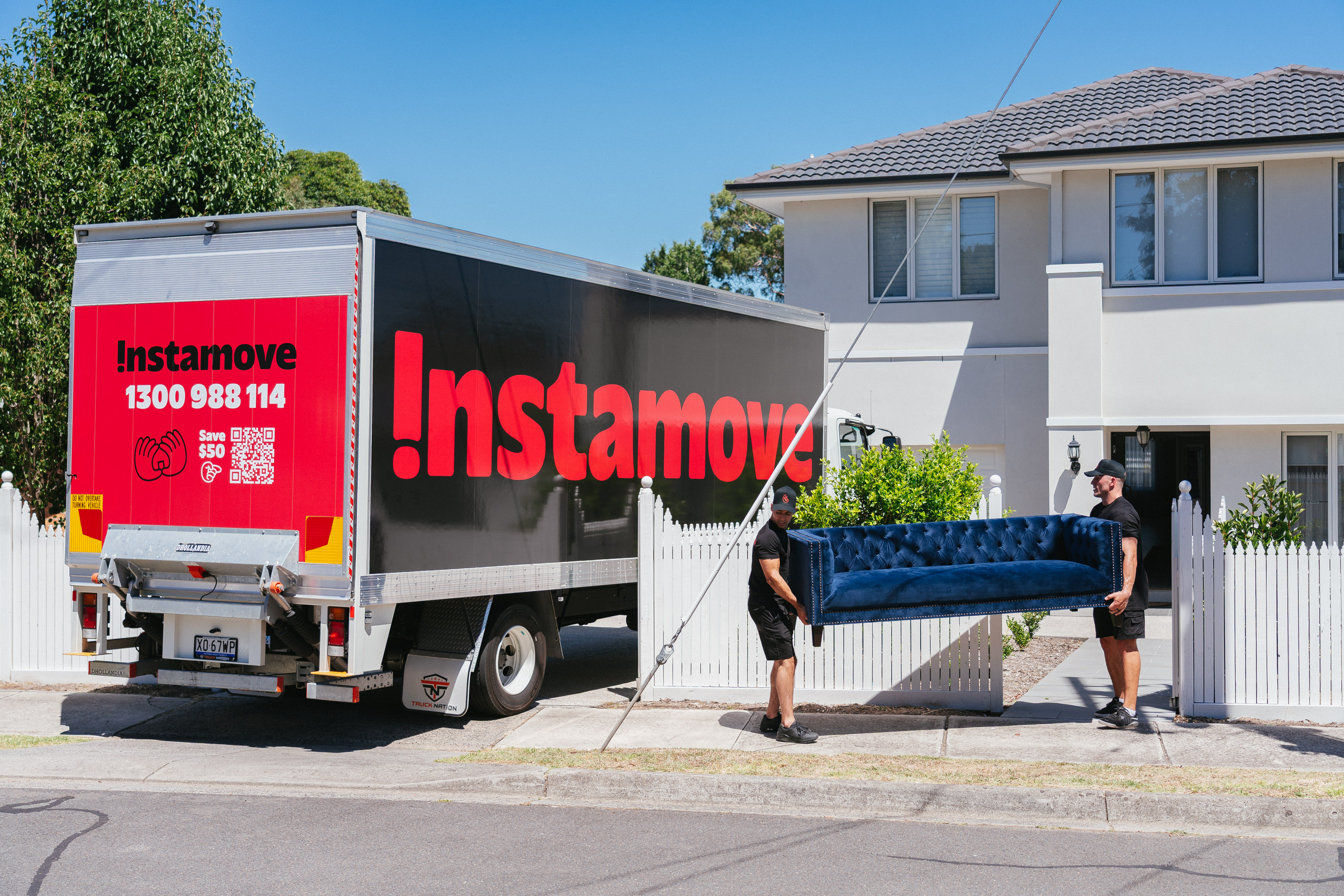 Instamove movers carrying a navy tufted sofa through a white picket fence gate with the branded truck parked on the street
