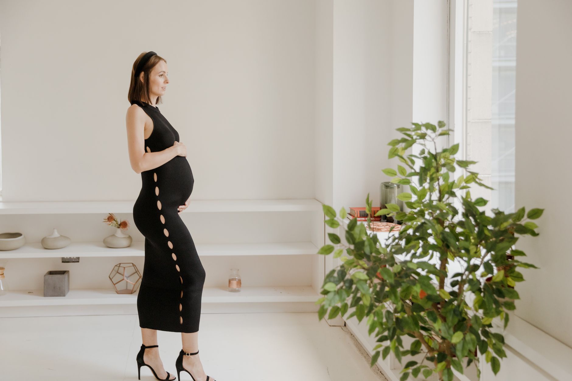 Pregnant woman wearing a soft, stretchy maternity dress, standing near a window with natural light, holding a coffee mug
