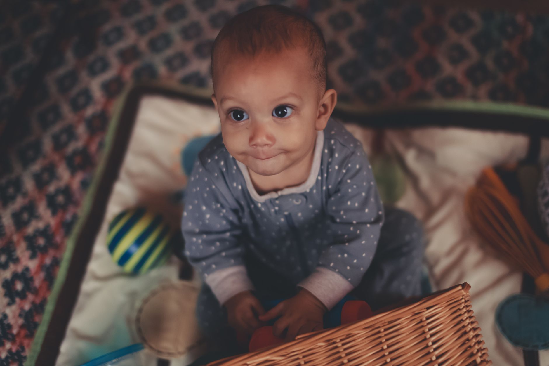 A smiling baby lying on a soft play mat, reaching toward a colorful toy while a caregiver watches nearby