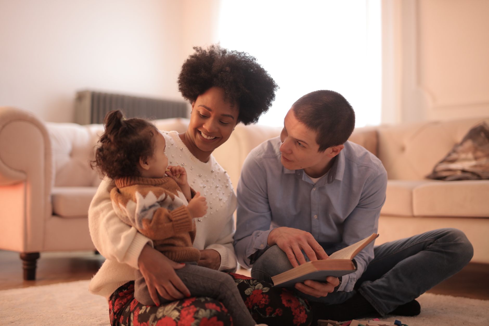 Family gathered on a living room rug, partner holding a baby book, older sibling drawing, soft natural light, warm tones