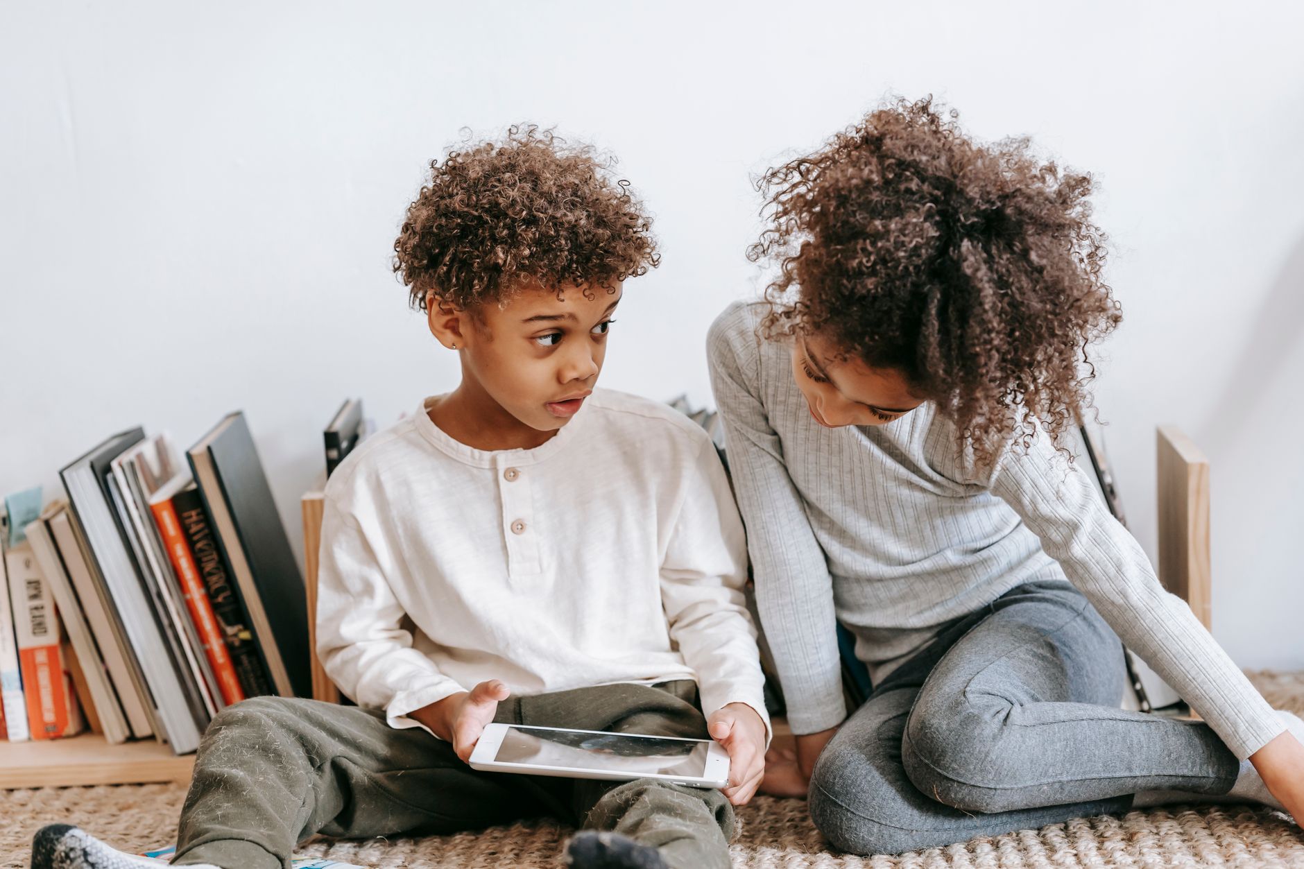 A parent sitting on the floor with a toddler and baby, giving each a snack while reading a book