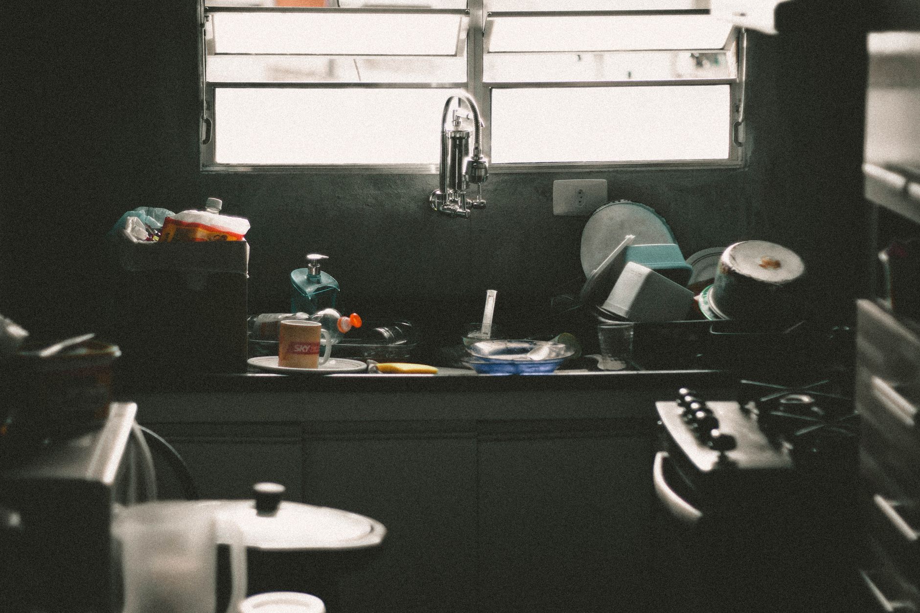 A chaotic kitchen counter with a baby monitor, coffee cup, and laptop showing a chore chart