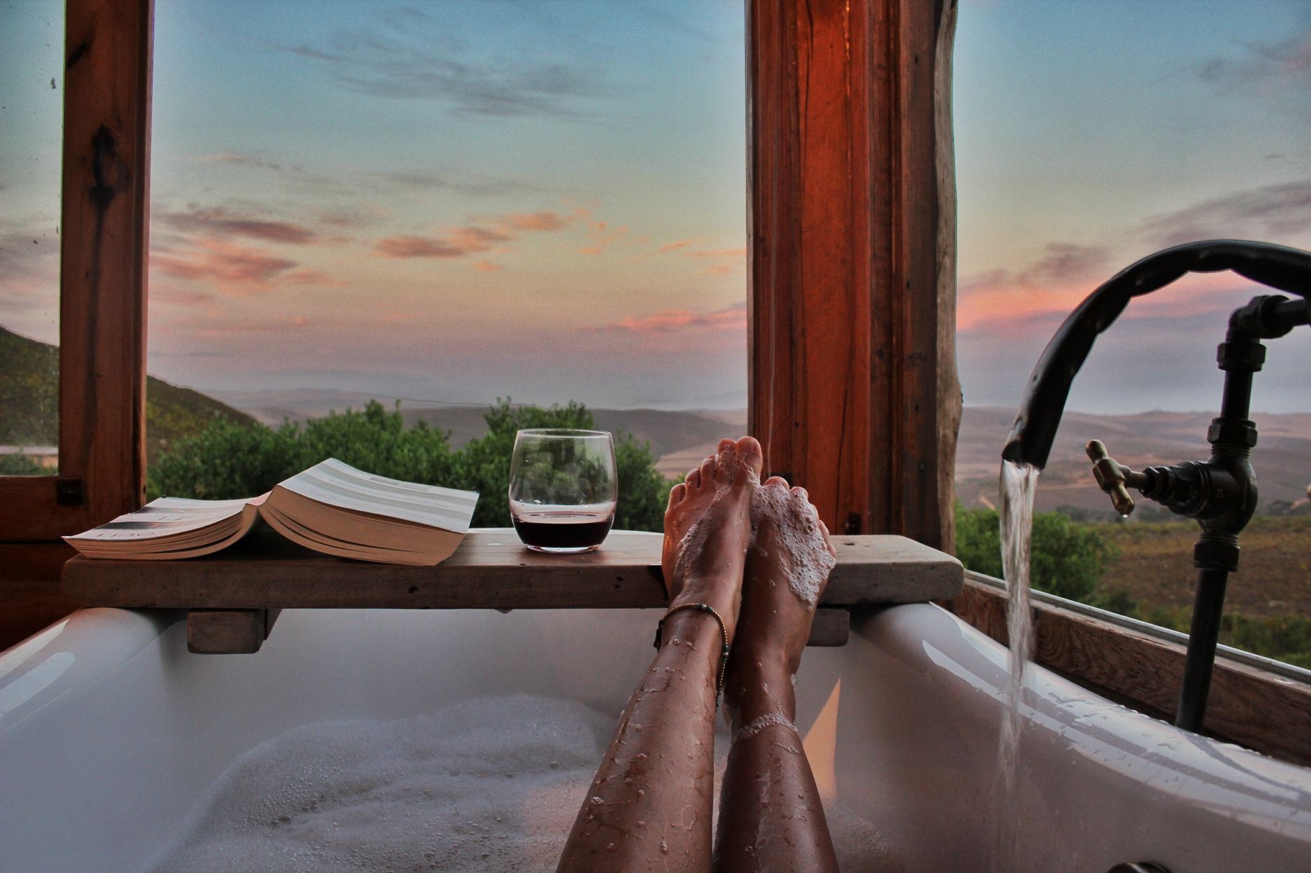A mom sitting in a warm bath with a book and a glass of wine, looking relaxed.