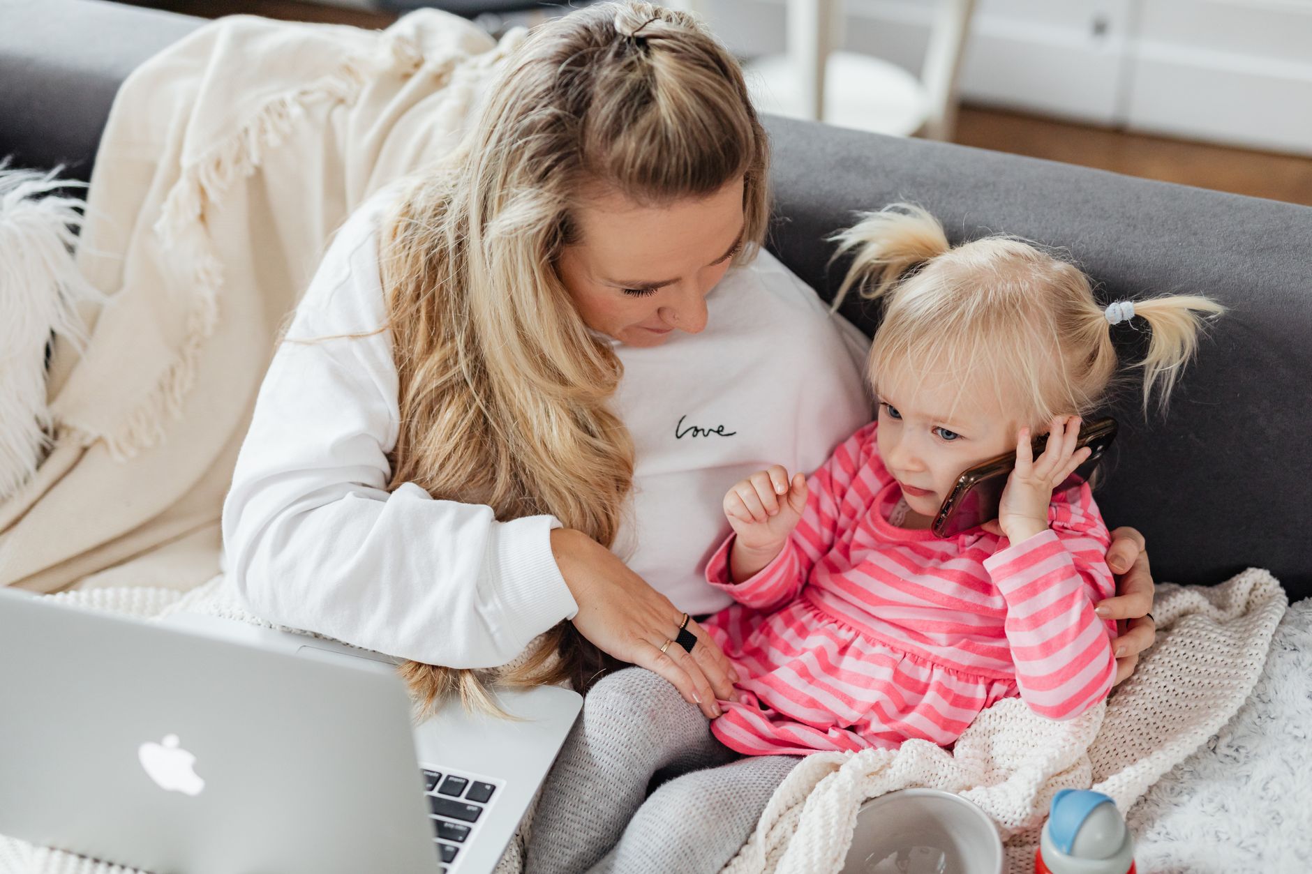 A mom smiling on her phone, holding a coffee cup, with her toddler playing nearby.