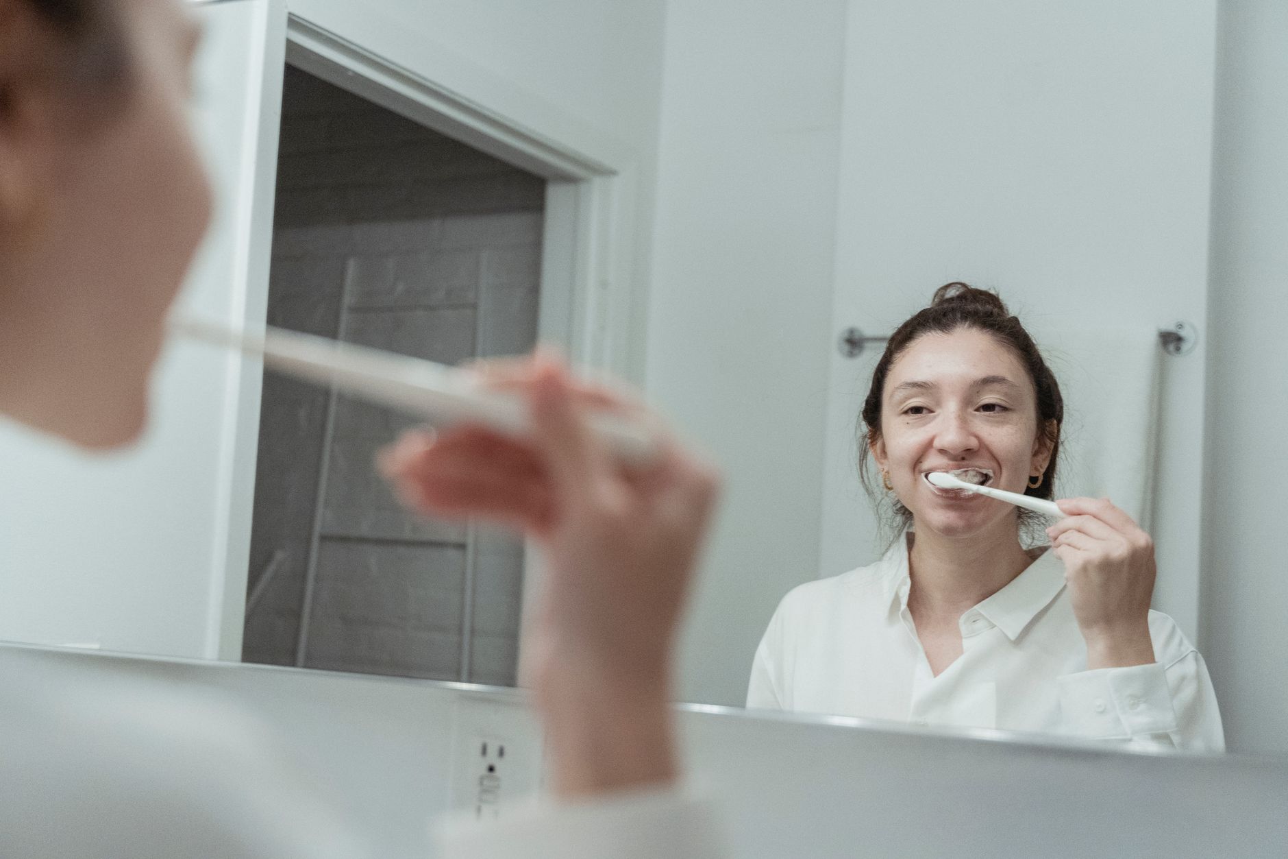 A pregnant woman smiling and holding a dental mirror, looking at her teeth, with a toothbrush in the background