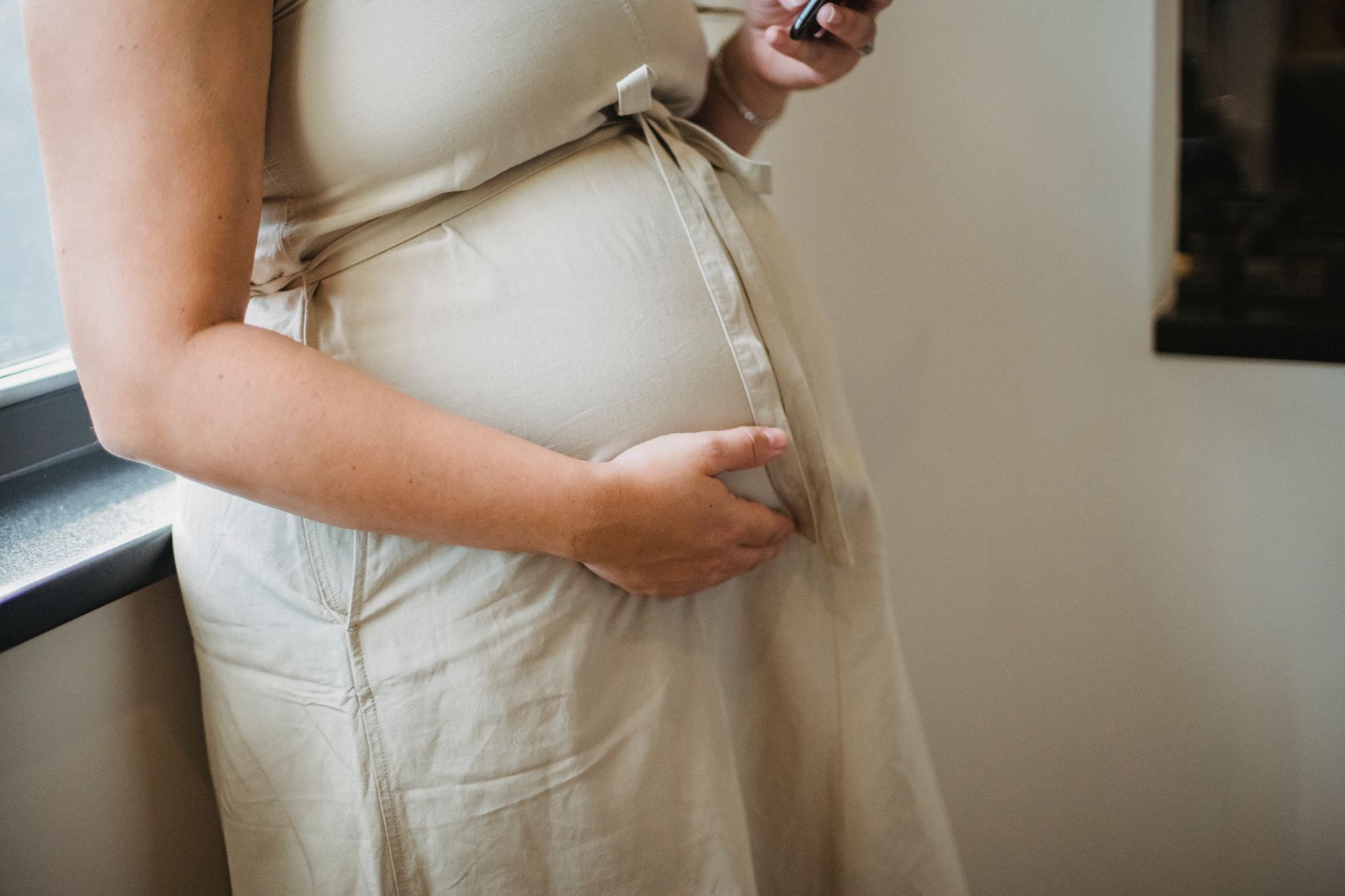 A pregnant woman gently placing a hand on her lower abdomen while sitting on a cozy couch, soft daylight streaming through a window, showing a relaxed, confident posture