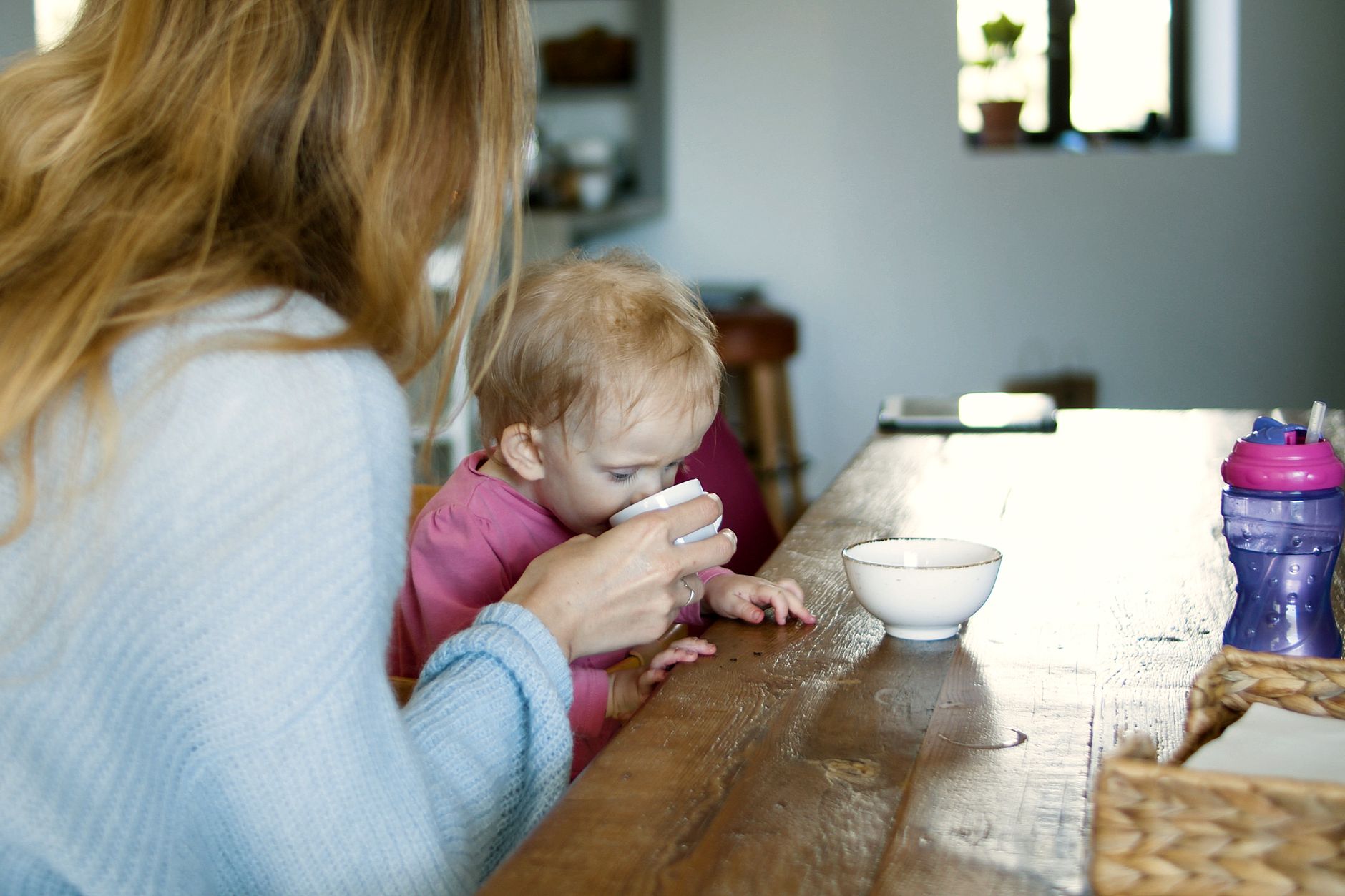 A mother and baby sharing a quiet moment on a sunlit balcony, with a bowl of fresh fruit and a water bottle nearby