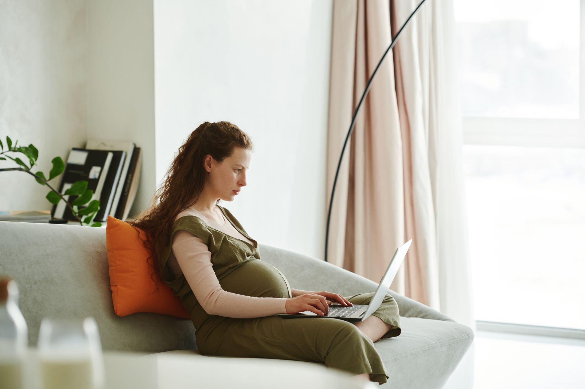 Pregnant woman holding a soft blanket, sitting on a couch with a laptop open, looking at a medical calculator page