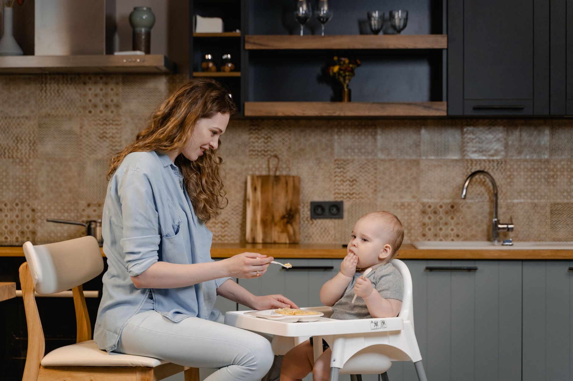 A mom and grandparent preparing baby food together in a warm, well-lit kitchen, with a jar of puree and a baby in a high chair.