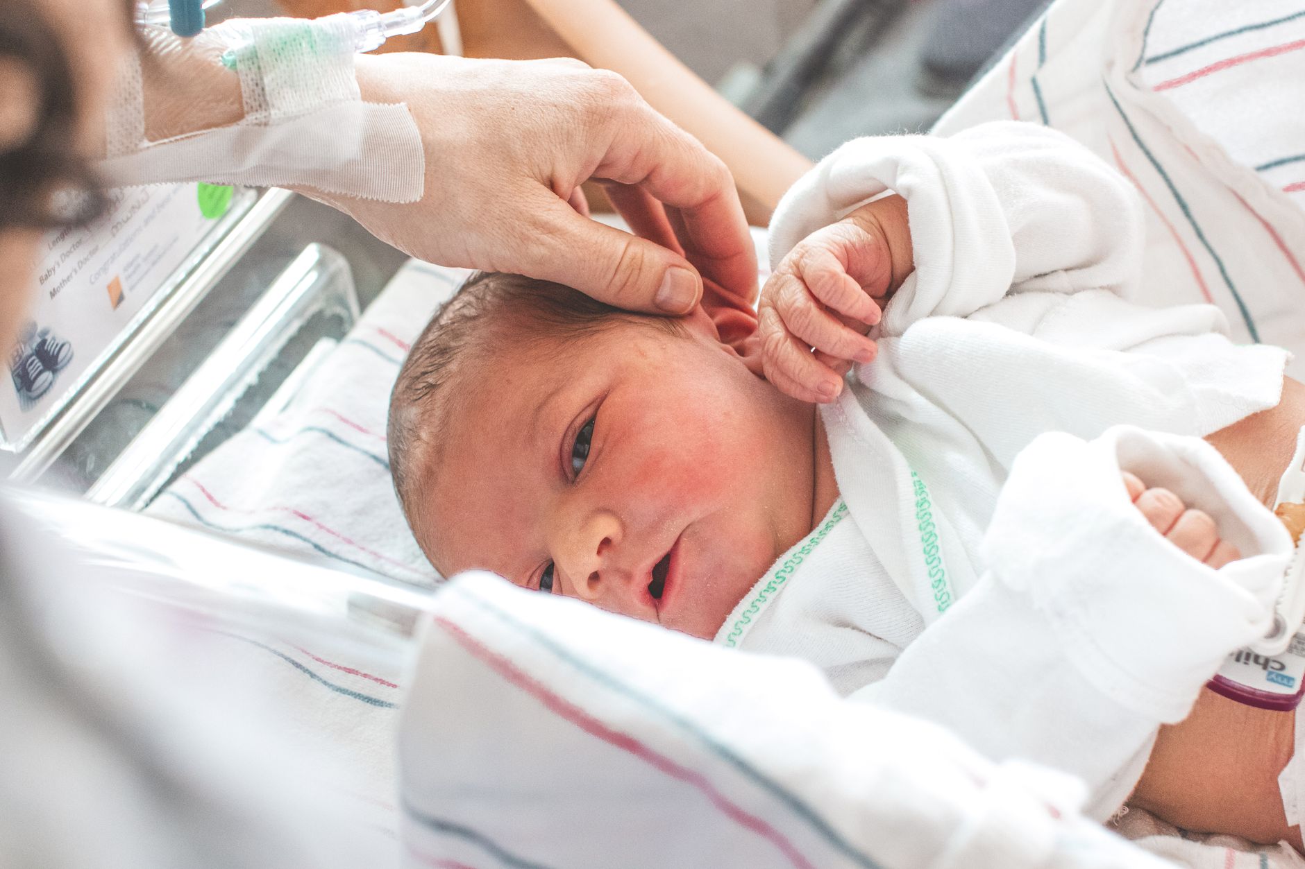 A child coloring at a small hospital table while a parent sits nearby, looking at a sleeping newborn.