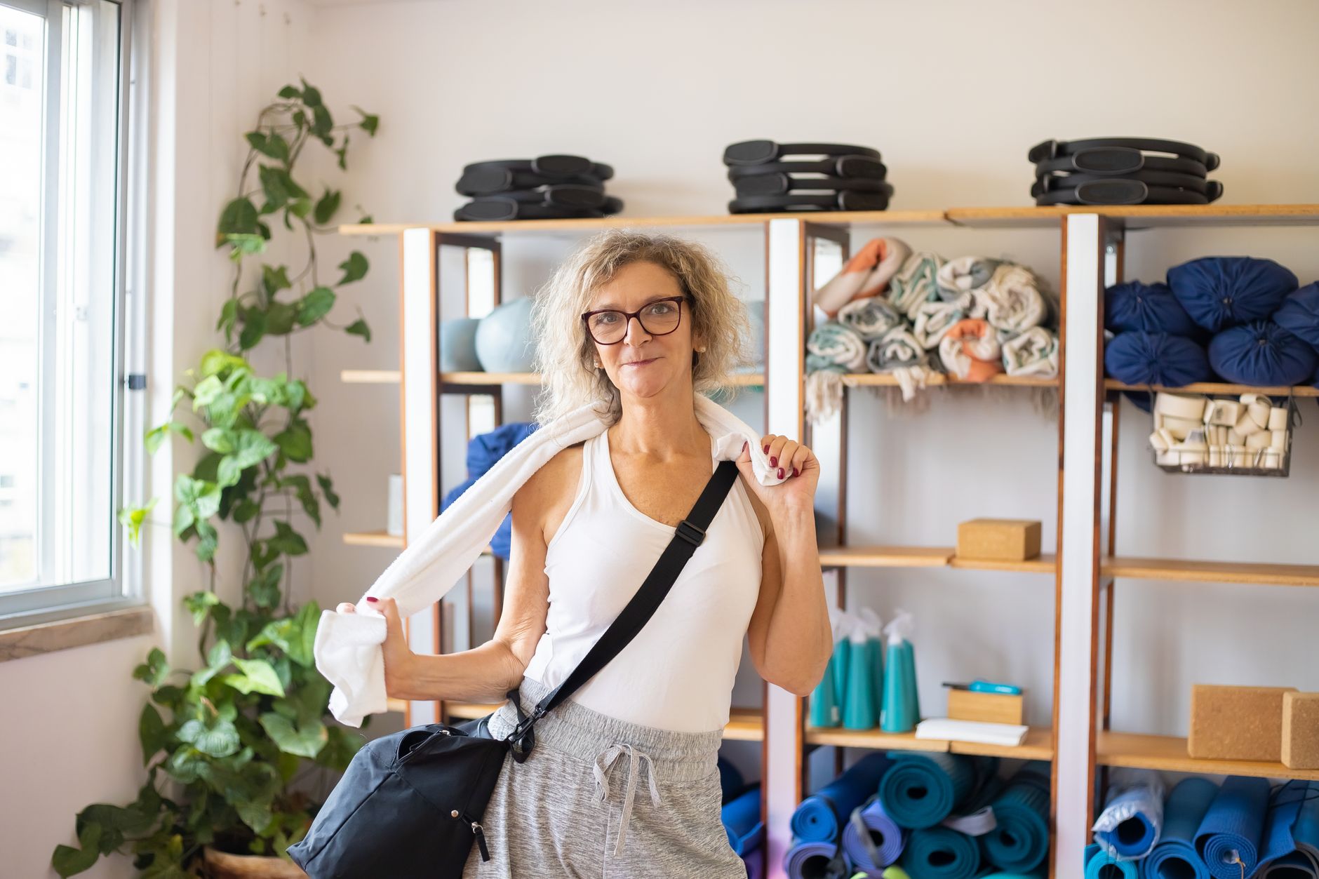 A mom holding a water bottle and a small towel, getting ready for hot yoga with a yoga mat and props in the background.