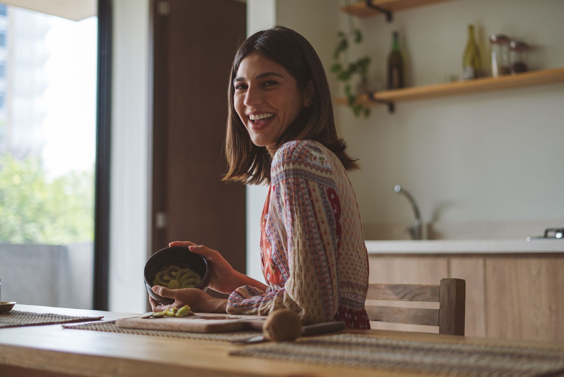 Pregnant woman holding a printed Chinese gender chart, smiling, with soft sunlight streaming through a kitchen window, fresh fruit on the countertop