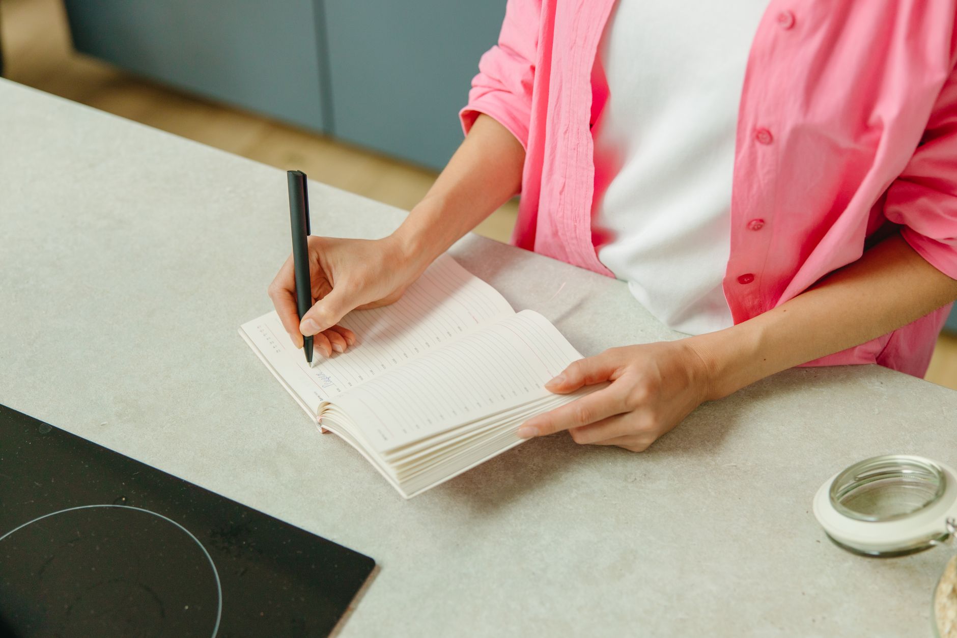 Soft-focus photo of a pregnant woman’s hands holding a small notebook with a pink calendar page marked, natural morning light, cozy kitchen table