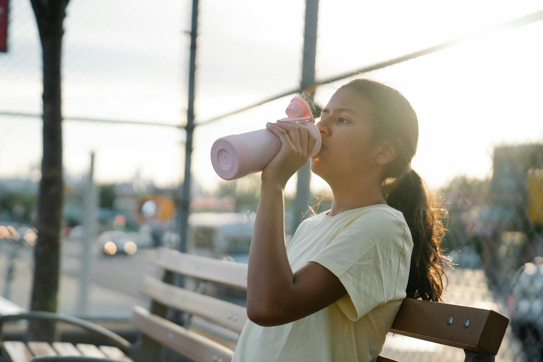 A pregnant woman holding a reusable water bottle while sitting on a sunny park bench, with a soft-focus background of green trees