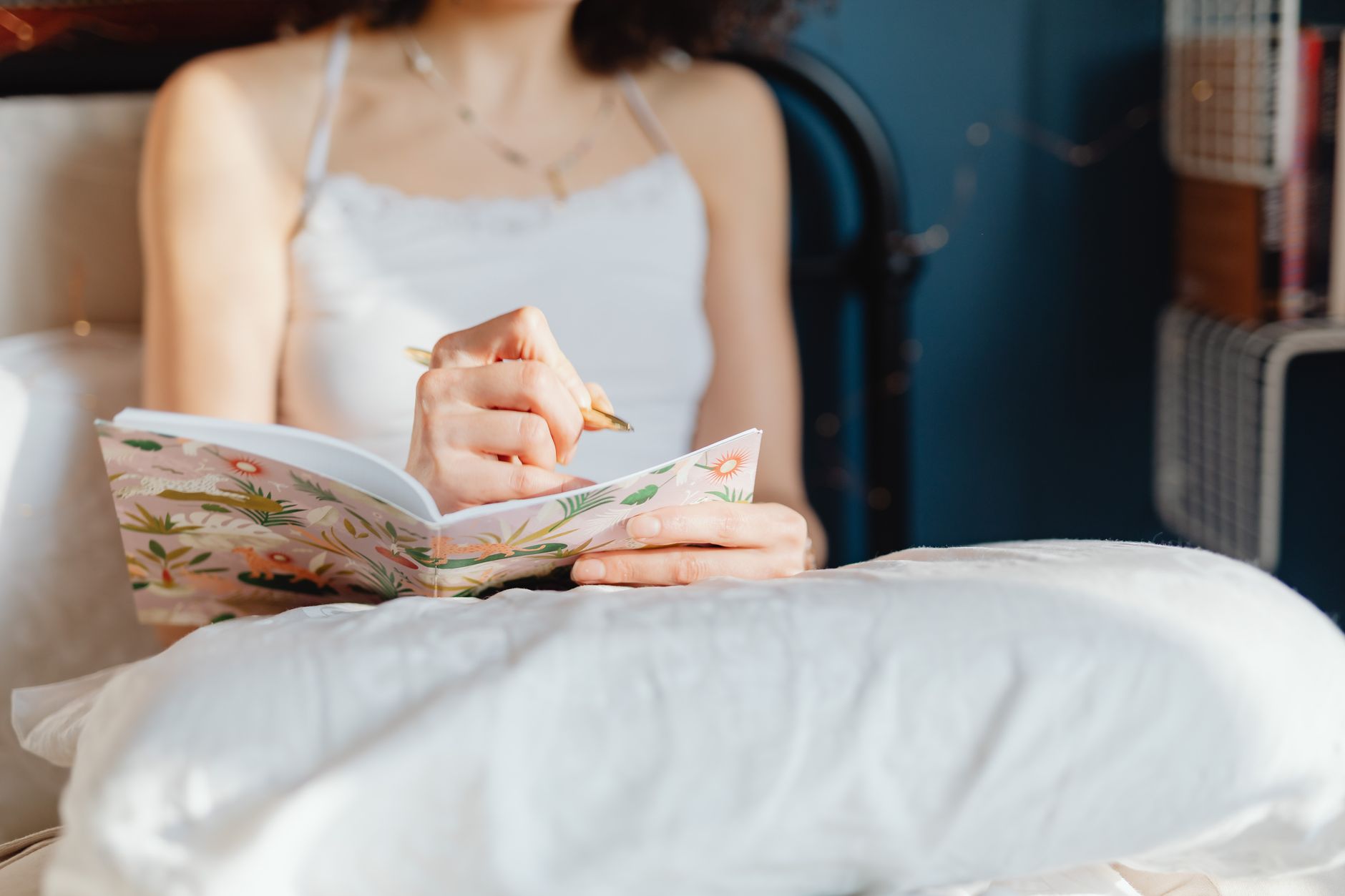 A woman holding a basal body thermometer next to a fertility chart with temperature readings, a pen, and a small notebook on a bedside table, soft morning light