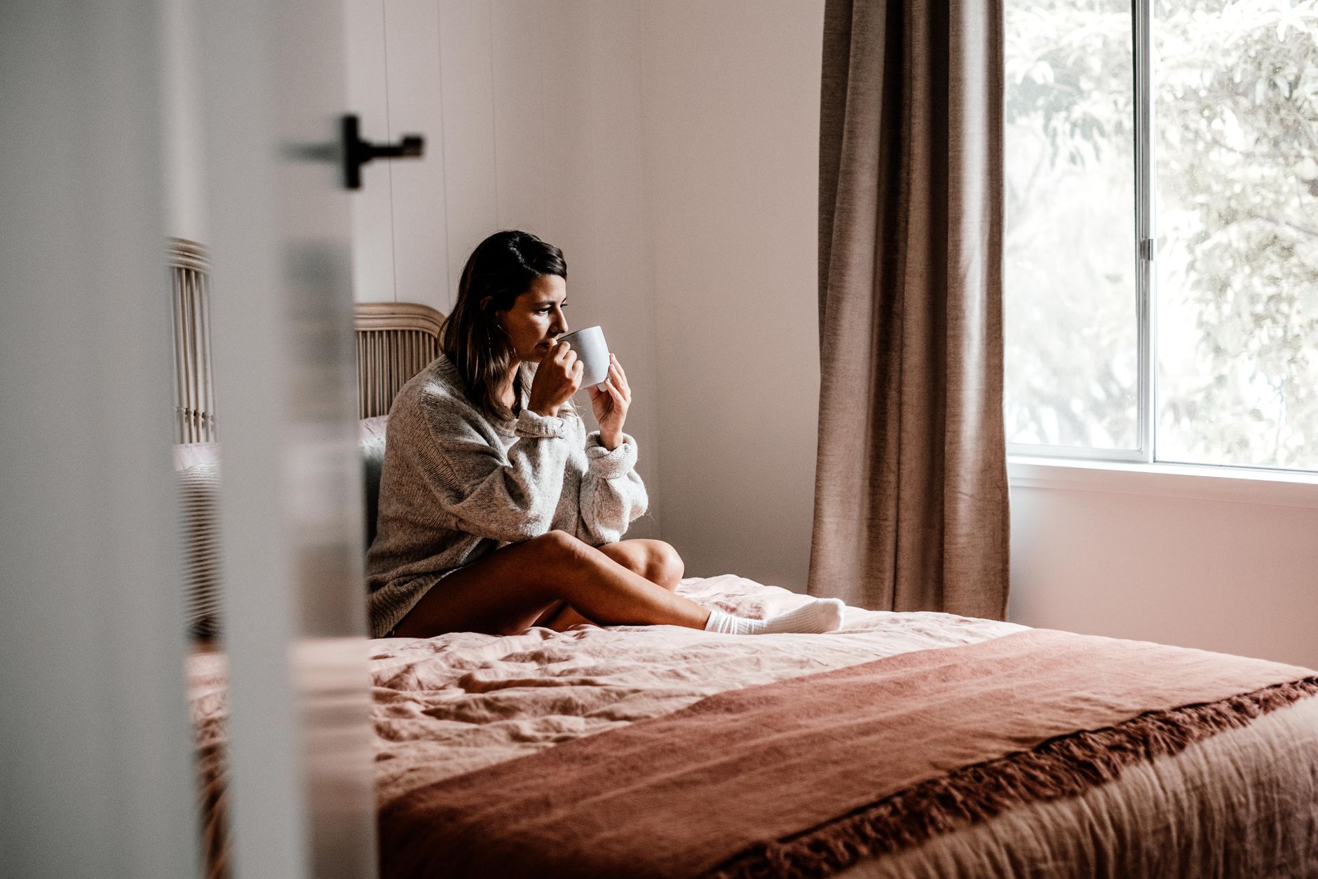 A serene bedroom scene with a pregnant woman sitting cross‑legged on a soft rug, sipping herbal tea, sunlight filtering through sheer curtains