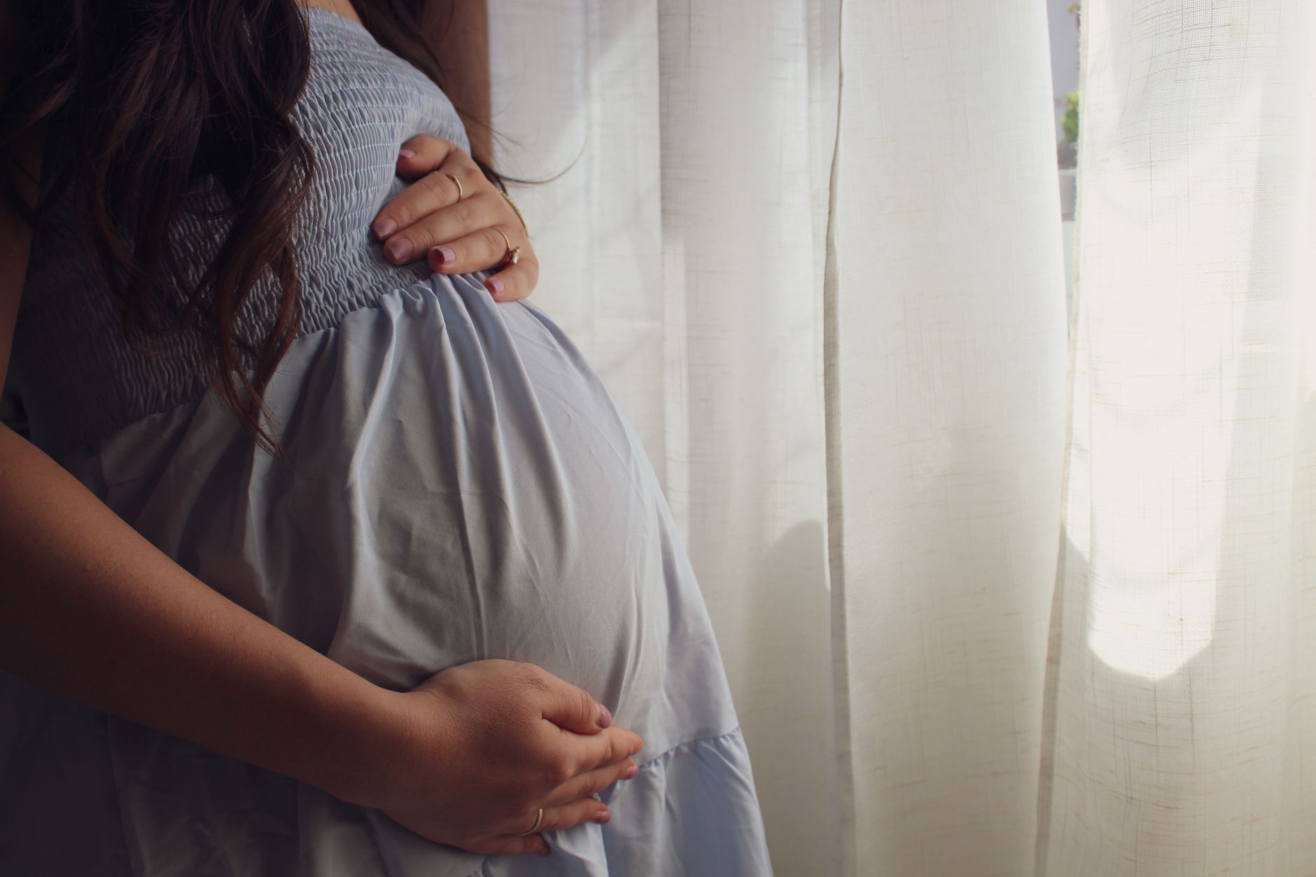 A pregnant woman gently touching her belly, looking out a window at a slightly hazy sky, with a serene expression.