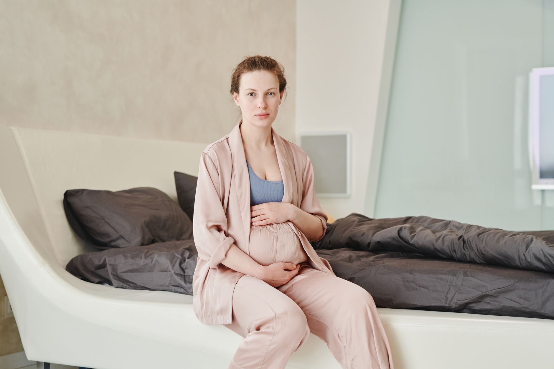 A pregnant woman's hand gently touching her belly, with a worried expression, sitting on a bed at night. Soft, warm lighting.