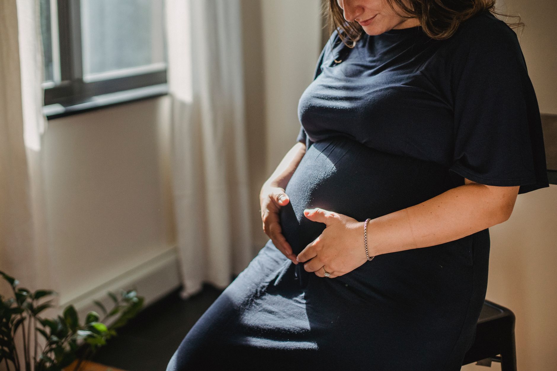 A pregnant woman gently touching her belly, sitting on a comfortable sofa, surrounded by soft blankets and a book, in a warm, cozy home environment.