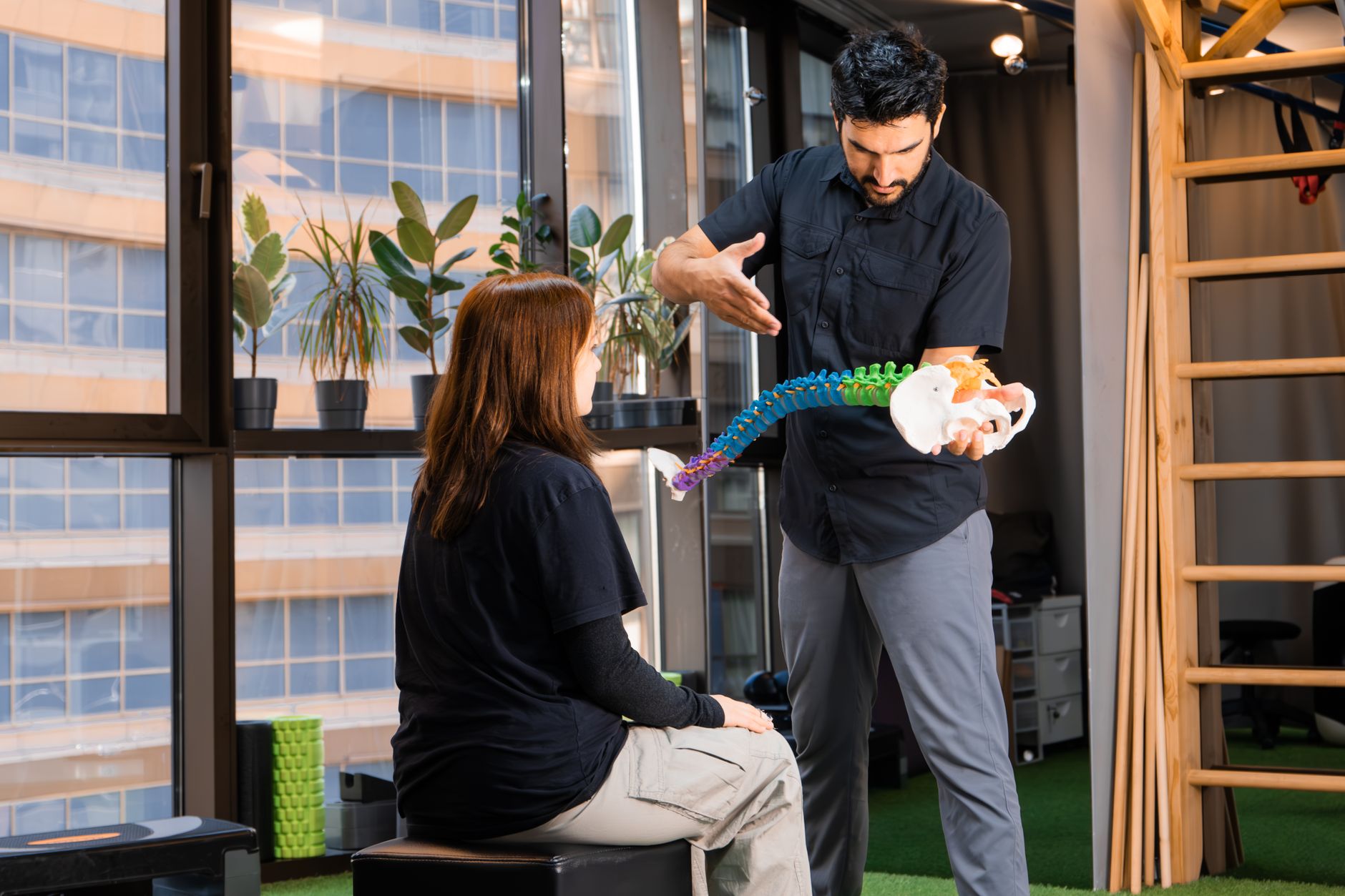 A prenatal chiropractor demonstrating the Activator Methods instrument to a pregnant woman, with a sign in the background listing the chiropractor's certifications, including Webster Technique and Activator Methods