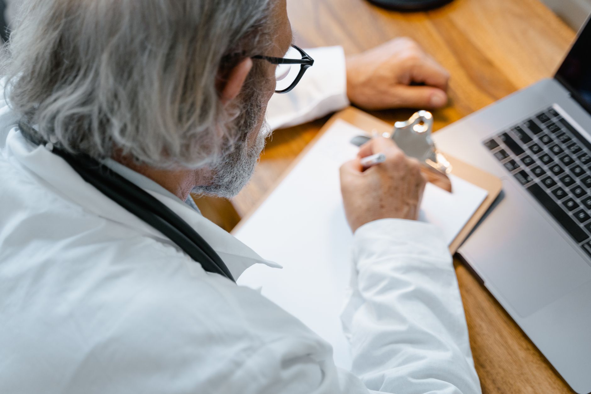 A medical professional, seen from the shoulders down, writing notes on a clipboard with a stethoscope nearby, in a clean, bright clinic setting