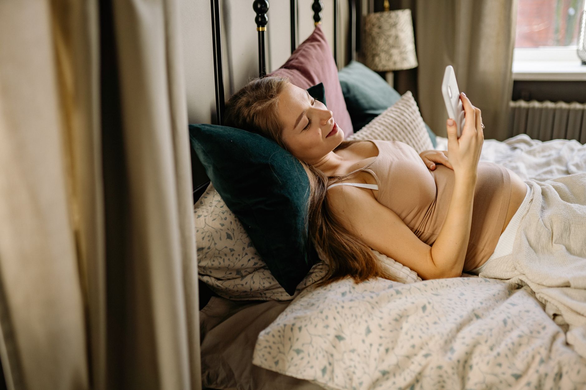 A pregnant woman talking on a smartphone, looking thoughtful and concerned, in a softly lit bedroom, with a pregnancy book on the nightstand