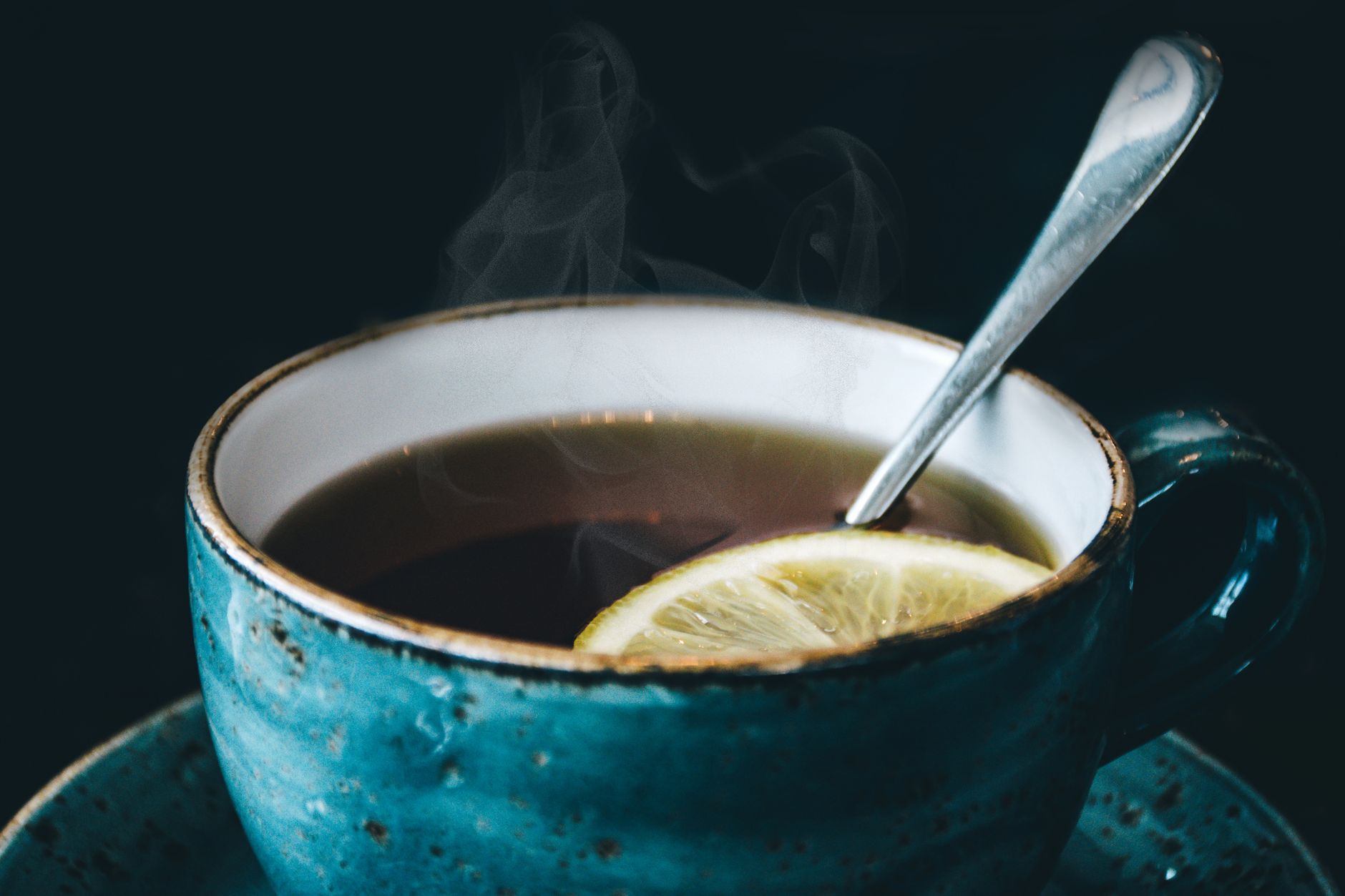 A steaming mug of honey-lemon tea with a slice of fresh ginger and a lemon wedge, sitting on a rustic wooden coaster.