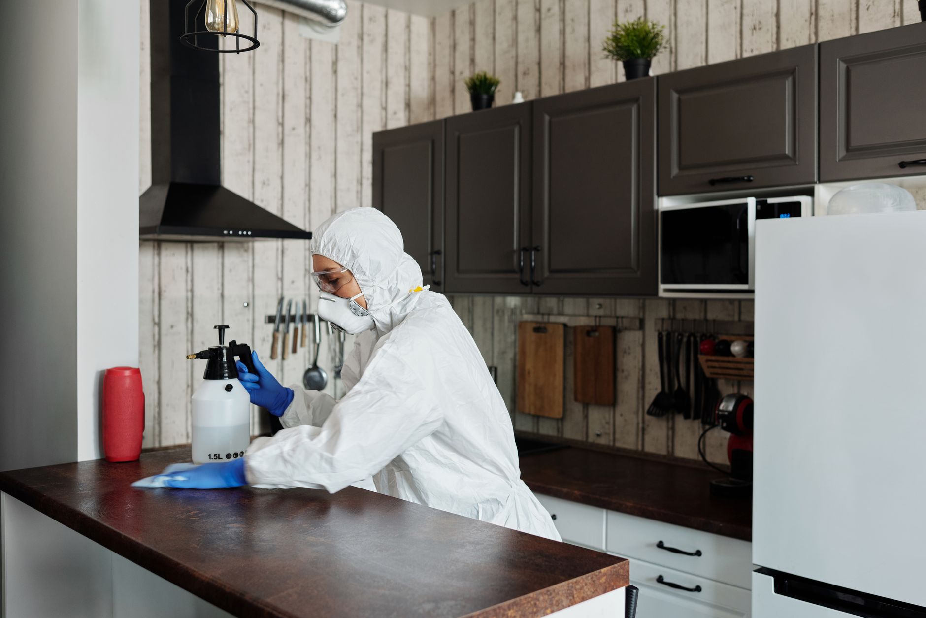 A pregnant person wearing gloves and a mask, cleaning a kitchen counter with a spray bottle and paper towels