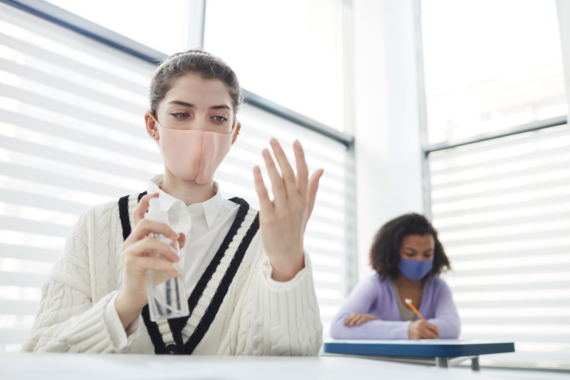 A pregnant woman at a school desk wearing a mask and using hand sanitizer while a child in the background plays with toys