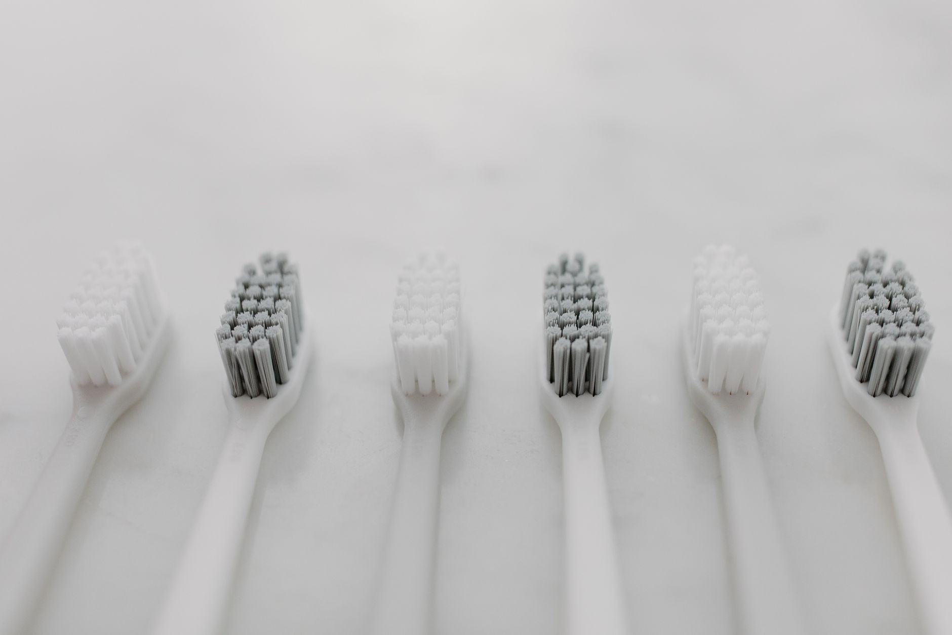 a row of baby oral‑care products on a light wood countertop, including fluoride toothpaste, a soft toothbrush, and a small cup of water, bright natural lighting