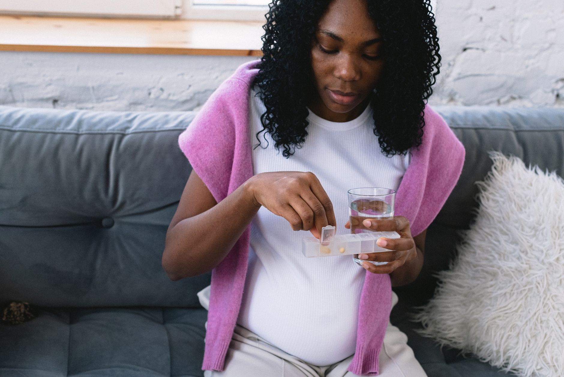 A set of prenatal vitamins, a glass of water, and a pregnancy journal on a clean, light-colored wooden nightstand. Soft, natural light from a window illuminates the scene.