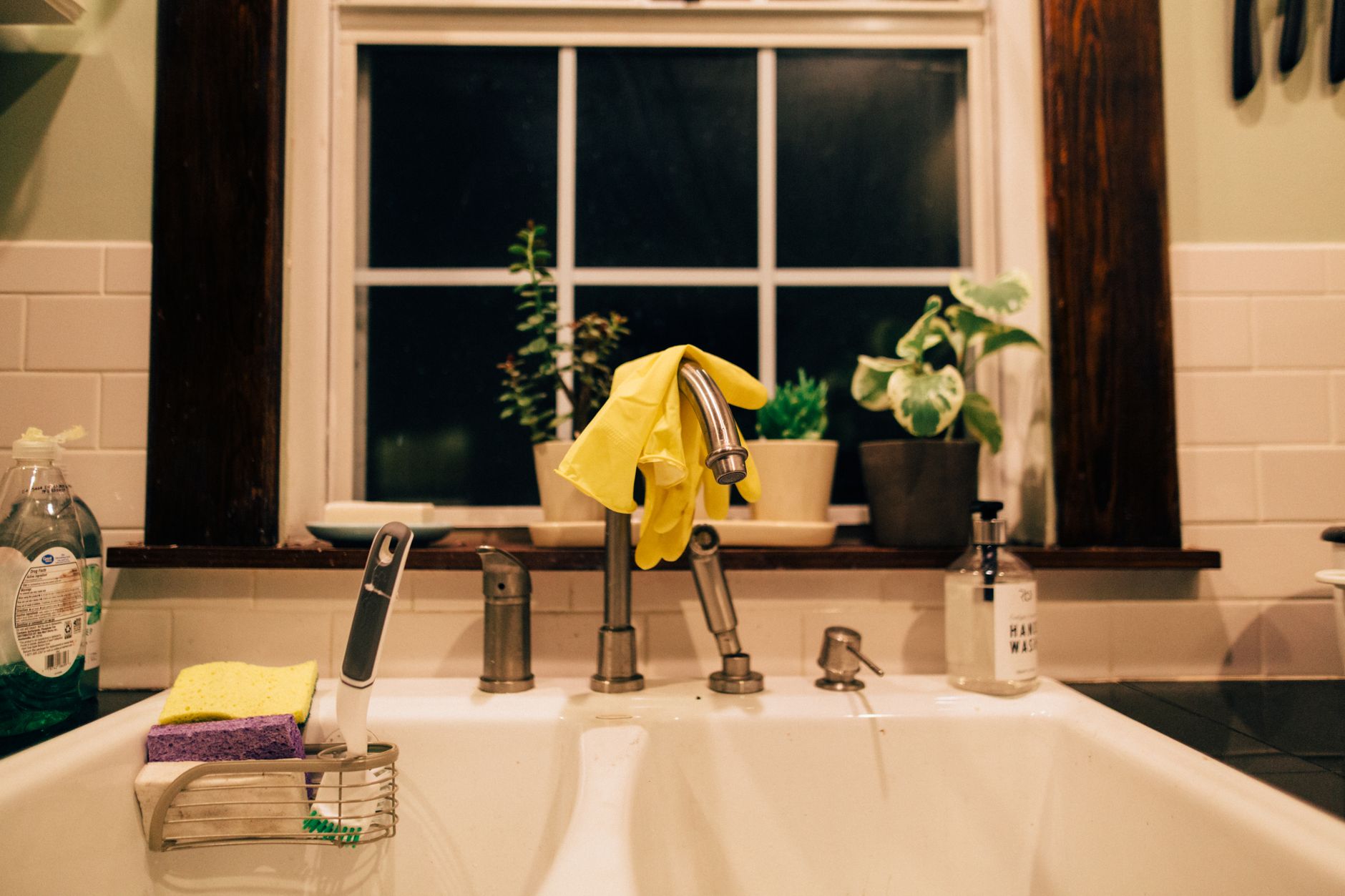 A pair of yellow rubber gloves and a green microfiber cloth resting on the edge of a clean kitchen sink, with a window providing natural light in the background.