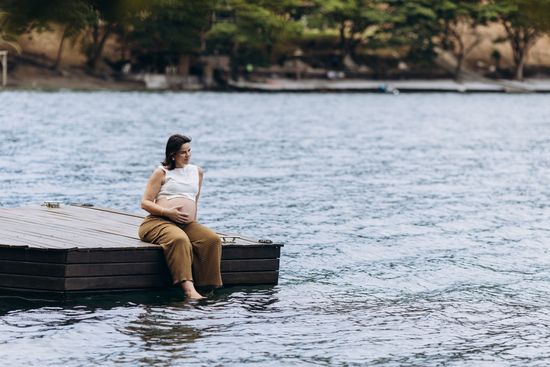 A pregnant person sitting on the shore of a calm lake, holding a paddle, with a serene landscape in the background