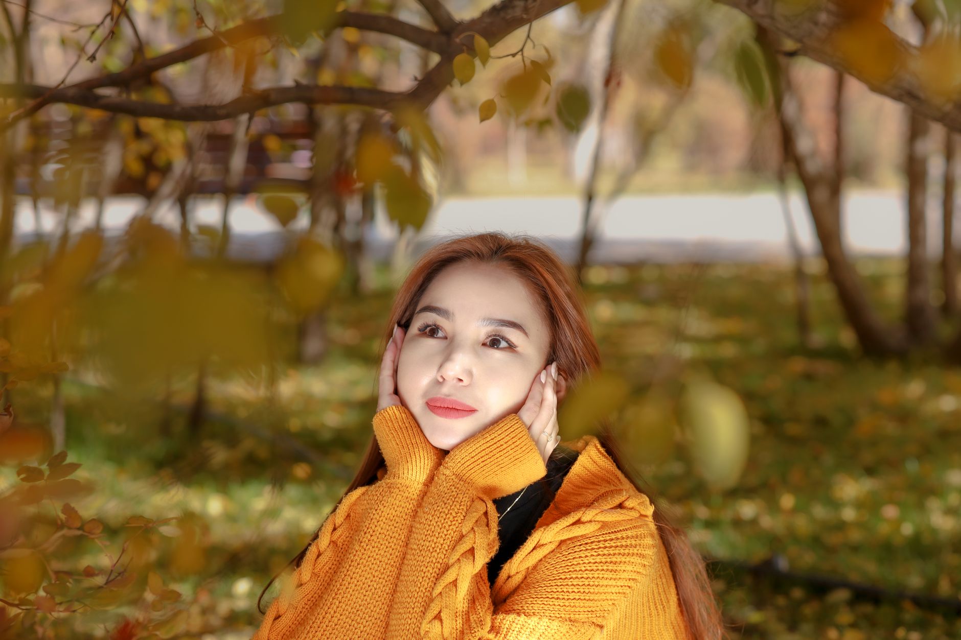 Pregnant woman wrapped in a soft knit cardigan, standing among autumn leaves with a gentle smile, warm light filtering through trees