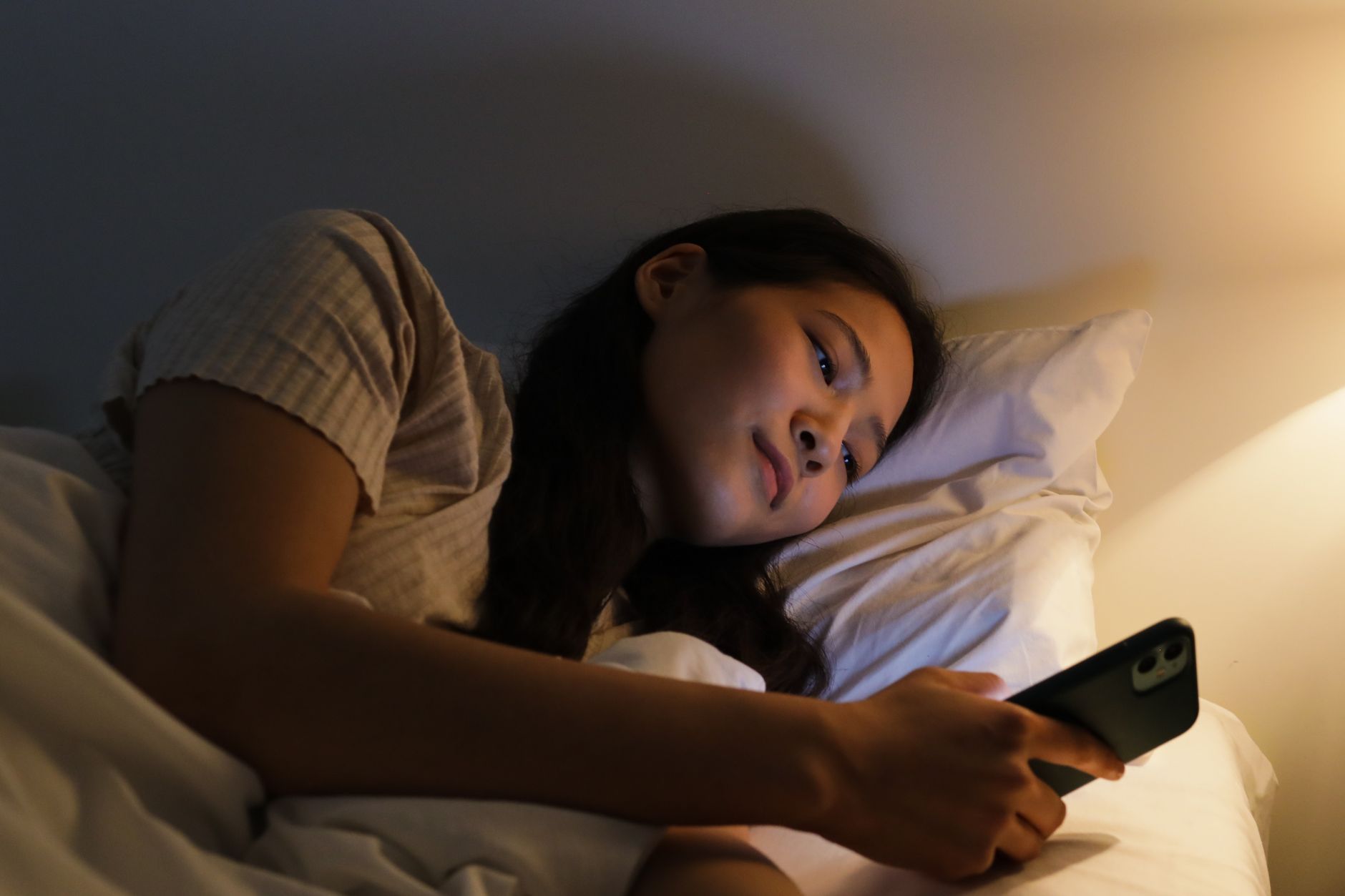 Woman lying in bed at night, holding a stuffed animal, looking at her phone.