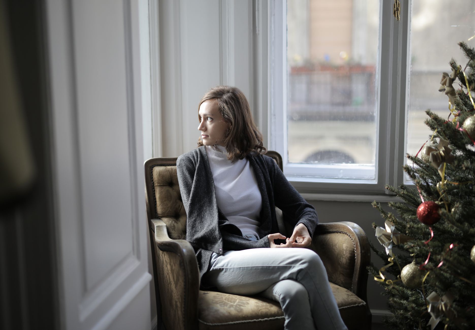 A woman sits thoughtfully on a sofa, looking out a window, with a warm, comforting atmosphere, representing reflection and support.