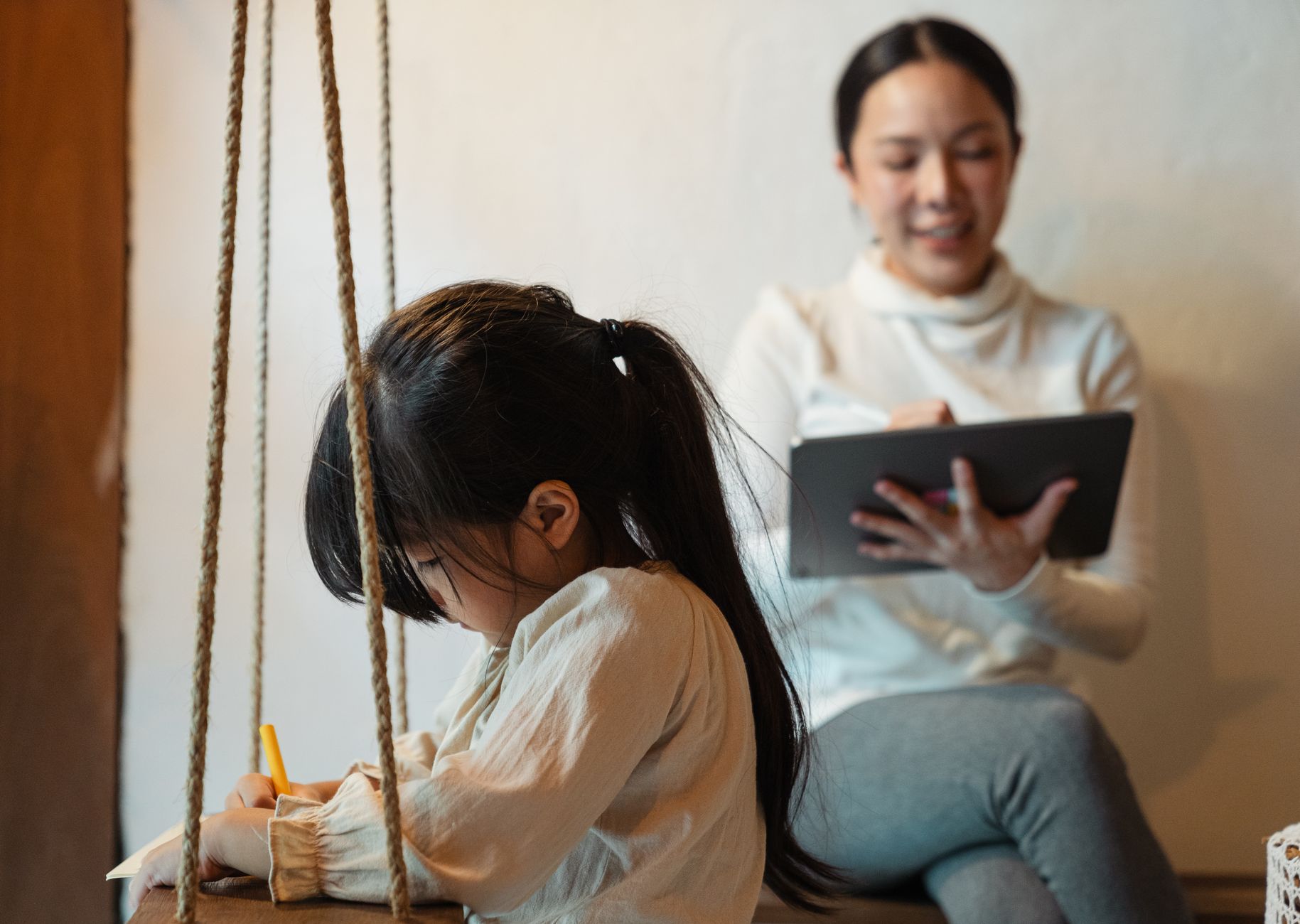 A woman in comfortable clothes sitting at a small table, writing in a notebook, with a child's toy visible on the floor in the background, illustrating a mom finding a moment to write amidst family life.