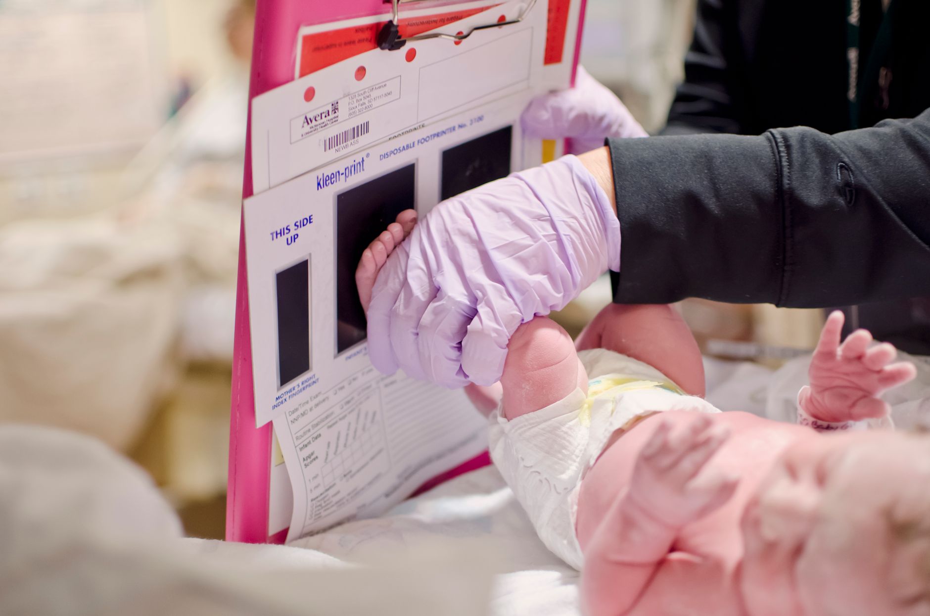 Close‑up of a nurse’s hand filling out a Finnegan scoring sheet beside a newborn’s crib