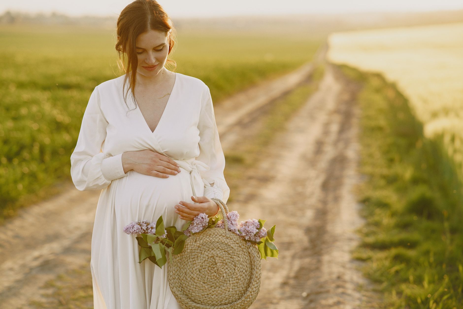 Pregnant woman standing in a sun‑lit meadow holding a flowing white dress, golden hour light creating a soft halo around her belly