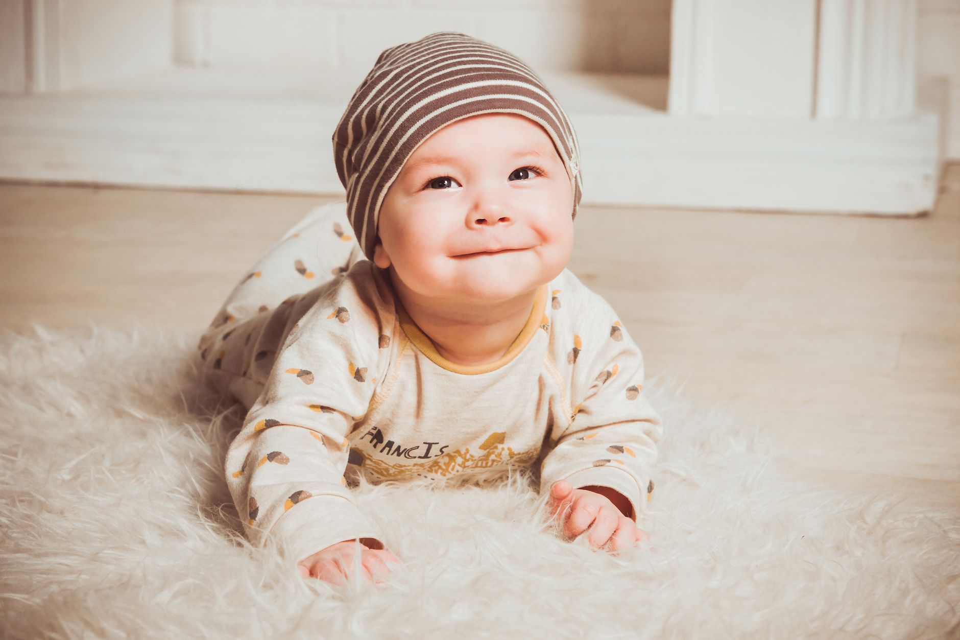 A baby in a light-colored cranial helmet, smiling and looking up at an adult's hand. The helmet appears comfortable and well-fitted.